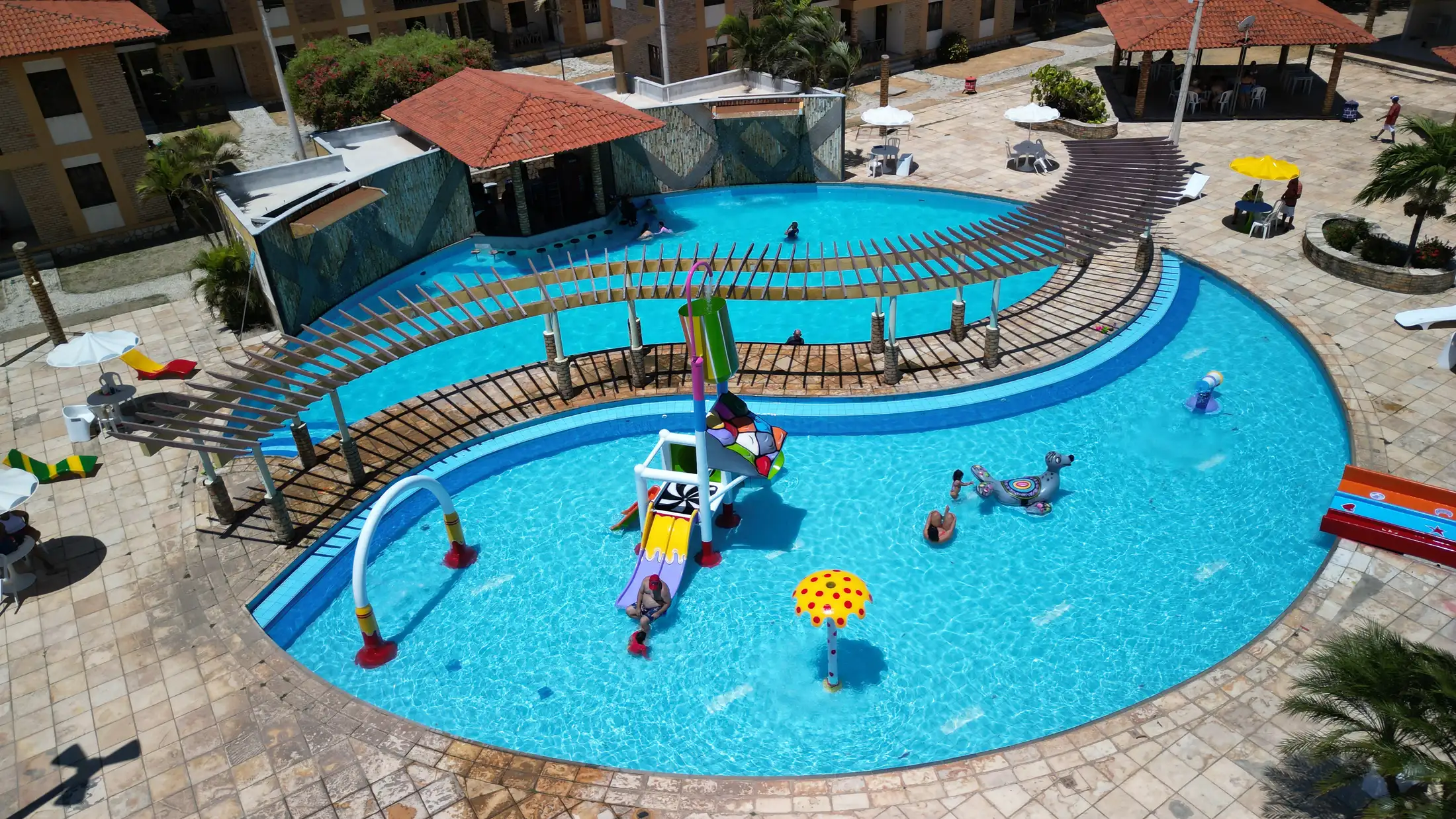 Vista aérea da piscina curva do Mirante das Gamboas, com pergolado de ripas de madeira e brinquedos aquáticos.